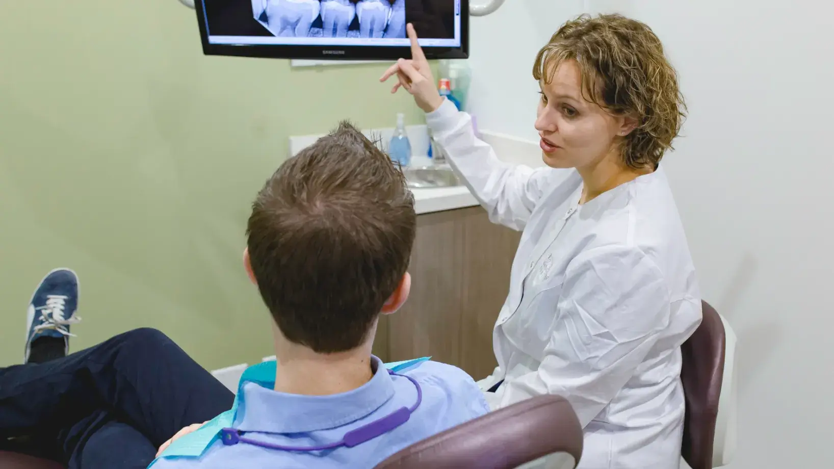 A woman in a dentist's office pointing at a man in a chair.